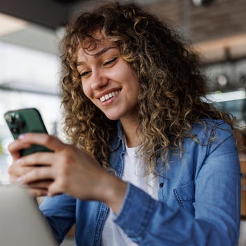 Young smiling woman using mobile phone while working on a laptop at a cafe